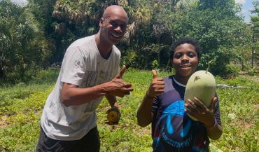 Two people giving thumbs up in a garden with a fruit.