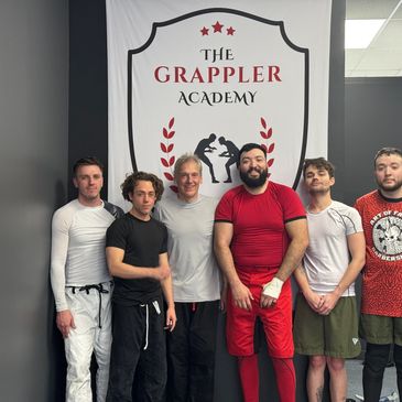 Six men posing in athletic wear in front of The Grappler Academy banner.