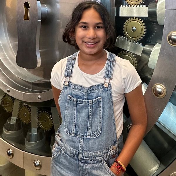 Girl in denim overalls smiles in front of a vault door.