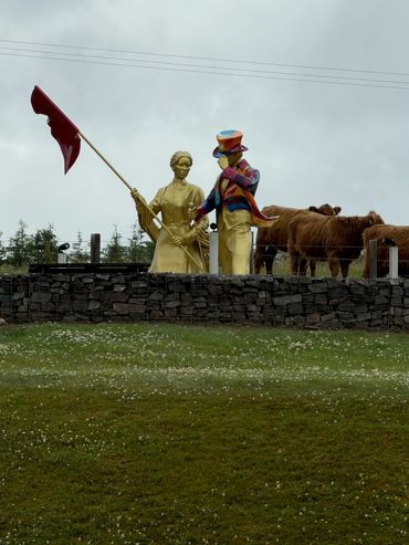 Golden statues of two figures near grazing cows on cloudy day.