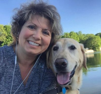 A woman happily posing with her smiling dog by a lake on a sunny day.