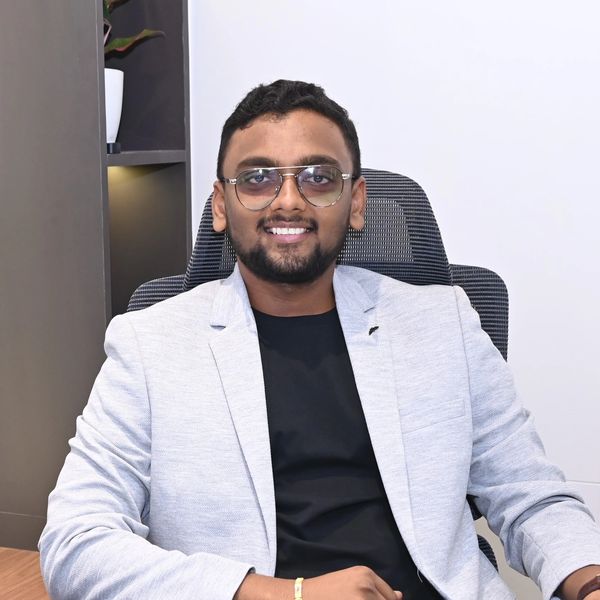 Man in light gray blazer and glasses smiling while seated.