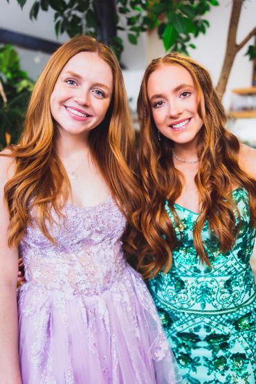 Two women with red hair smile at the camera wearing purple and green dresses. 