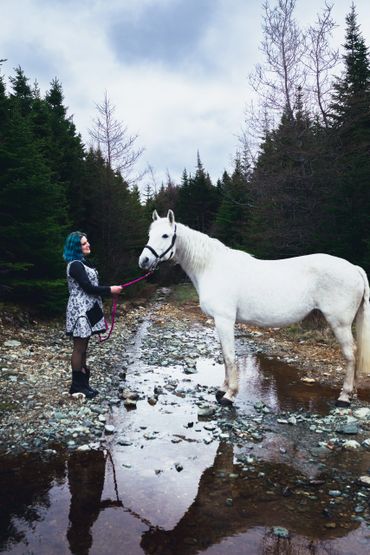 Woman with turquoise hair stands facing a white horse. Their reflection is showing in a puddle. 