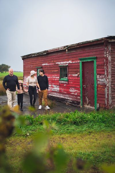 Family of four walks holding hands in a grassy scene in front of an old red shed. 