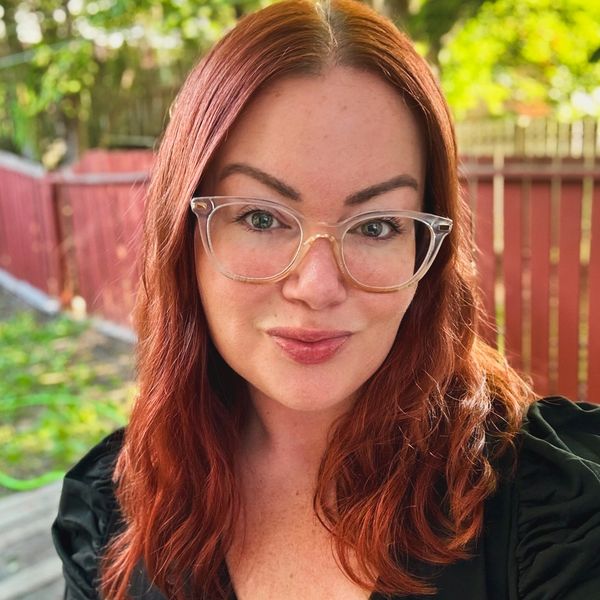 A woman with red hair and clear glasses smiling outdoors.