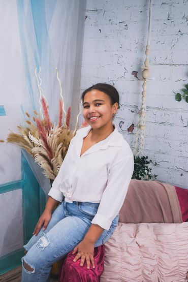 Girl with black hair sits on a pink chair and smiles. 