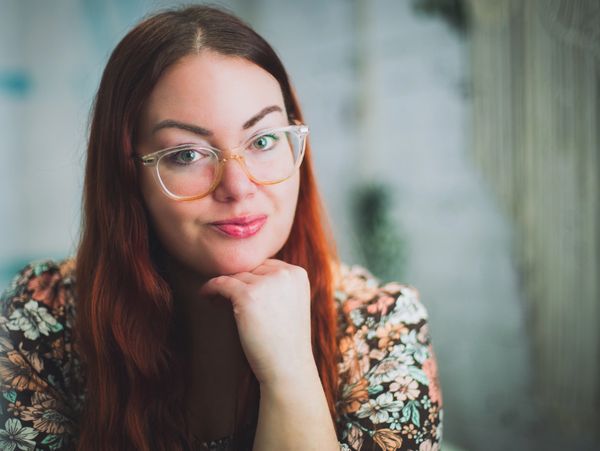 Woman with red hair and clear framed glasses smiles at the camera with her hand under chin. 