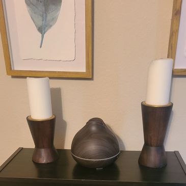 Two candles in wooden holders and a diffuser on a black table under framed art.