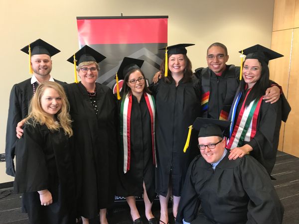Kristopher Oliveira with his Masters cohort at their hooding ceremony. All are wearing graduation an