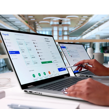 Person working on payroll and email management on two laptops in a modern office.