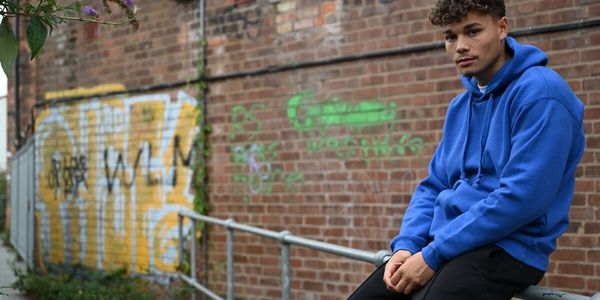 Young man in blue hoodie sitting on a railing against a graffiti-covered brick wall.