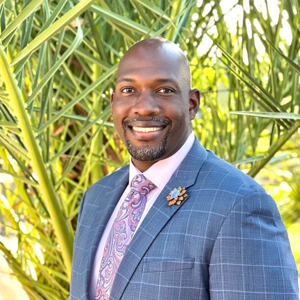 Smiling man in a blue checked suit with a paisley tie, standing outdoors near green palm leaves.