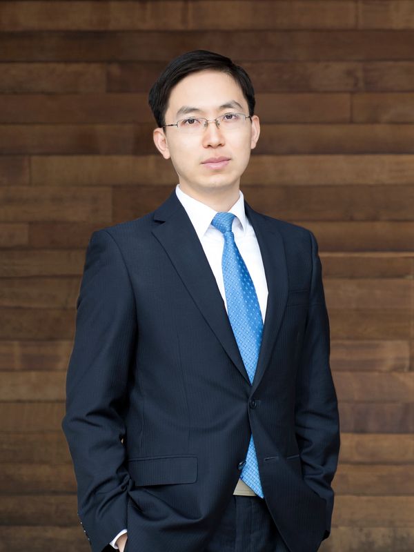 Professional man in a dark suit and blue tie standing against a wooden wall.