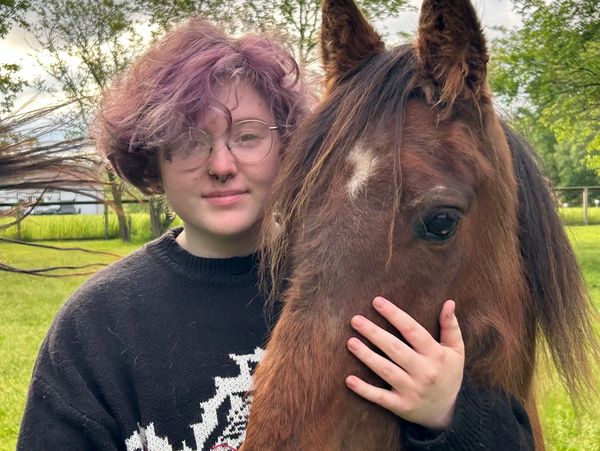 Person with glasses and purple hair hugging a brown horse outdoors.
