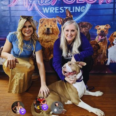 Two women smiling with a dog in front of a colorful wrestling-themed backdrop.