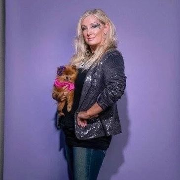 Woman in sequin jacket holding a small dog against a purple backdrop.