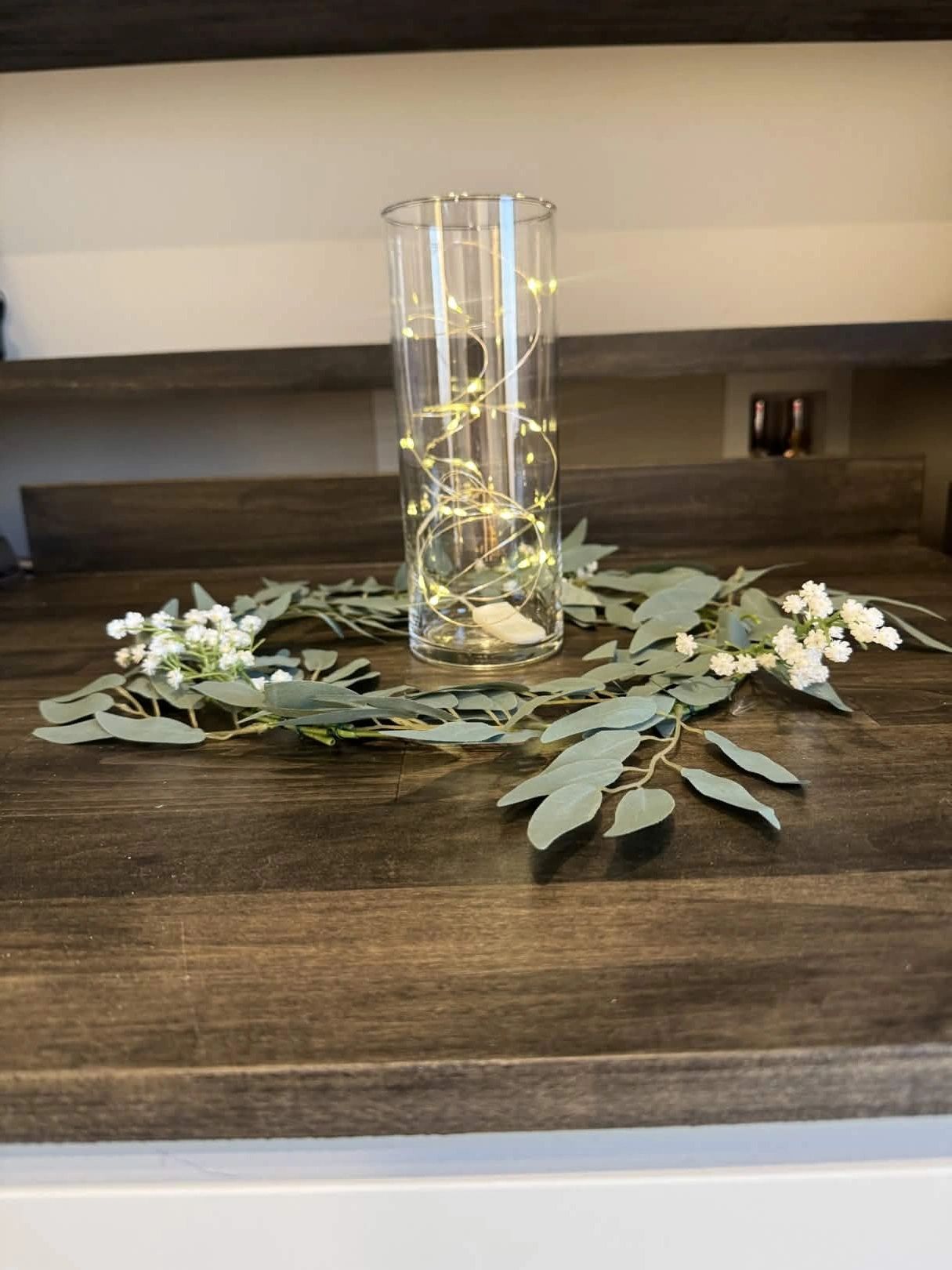 Glass vase with string lights surrounded by green leaves and white flowers on wooden surface.