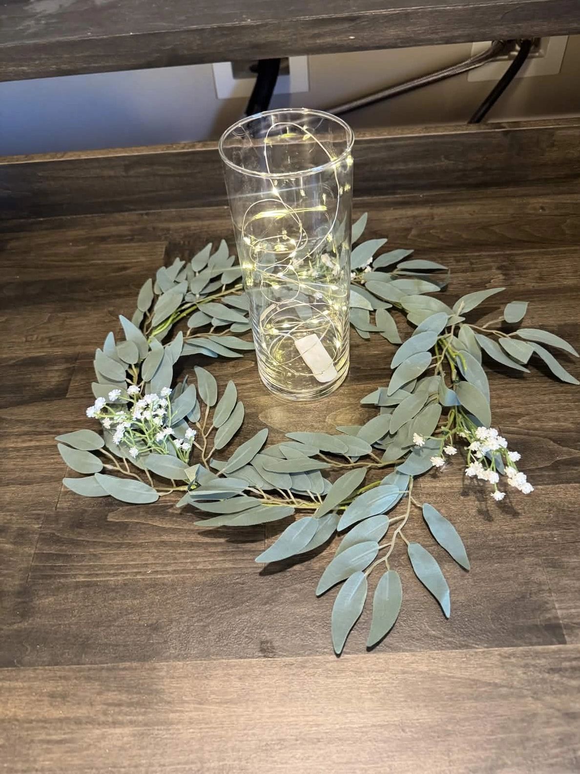 A glass vase with fairy lights surrounded by a green leafy wreath with small white flowers.
