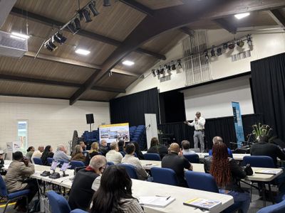 A speaker addressing an audience in a conference room with blue chairs.