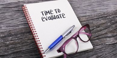 A notebook with time to evaluate written on the front, a pen and a pair of spectacles