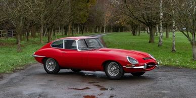 Classic red Jaguar E-Type parked on a tree-lined driveway.