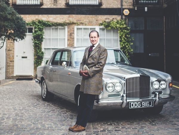 Man in a coat posing beside a classic Rolls-Royce car on a cobblestone street.