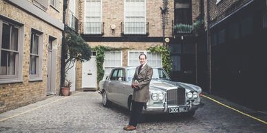 Man standing confidently in front of a classic silver car in a cobblestone alley.