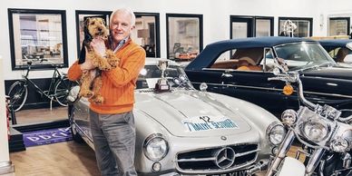 Man holding a dog in a vintage car showroom with classic Mercedes and motorcycle.