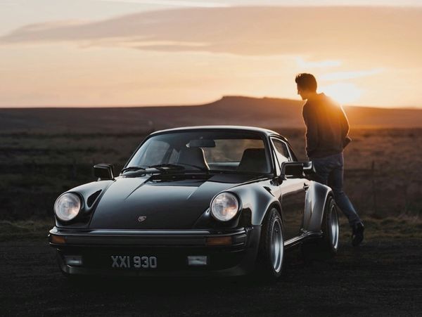A classic black Porsche 911 with a person standing beside it during sunset.