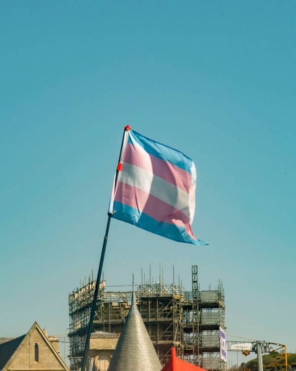 Transgender pride flag waving against a clear sky above a building under construction.