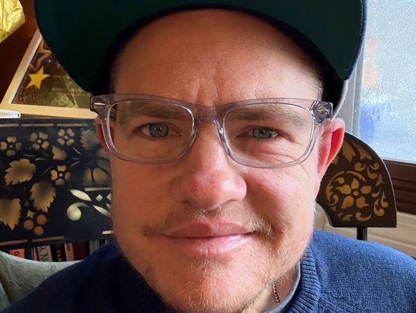 Close-up of a smiling man wearing clear glasses and a cap.