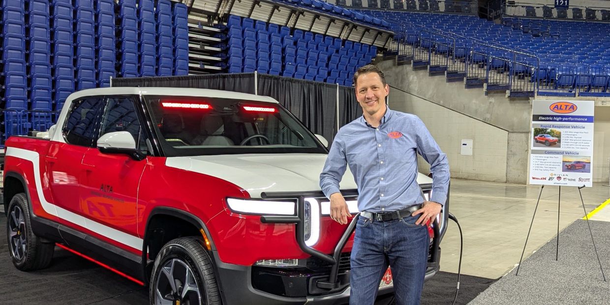 Man posing beside a red and white electric truck at an indoor event.