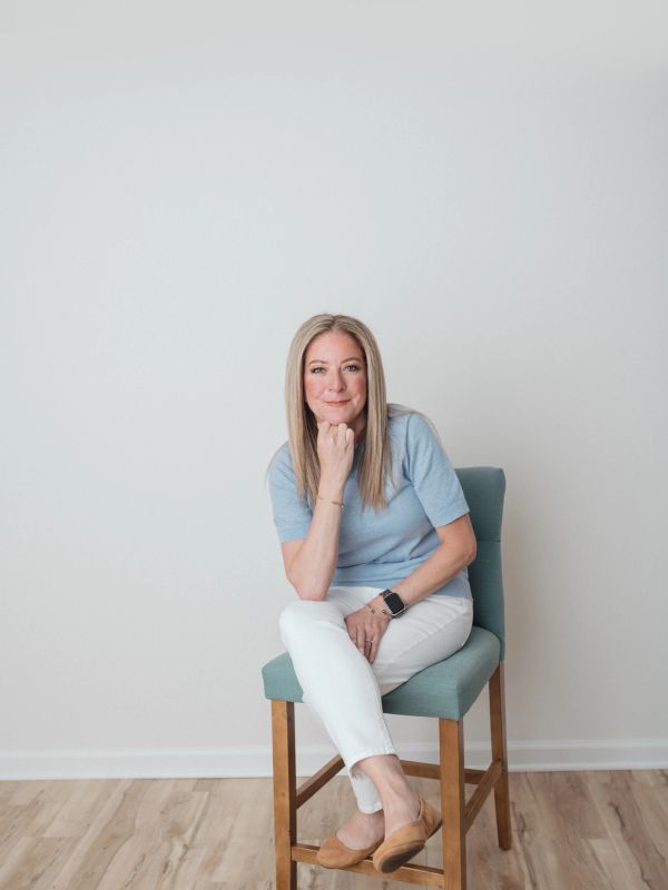 Woman sitting on a wooden chair against a plain wall.