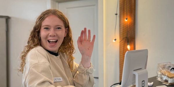 Smiling woman waving while working at a point-of-sale terminal.