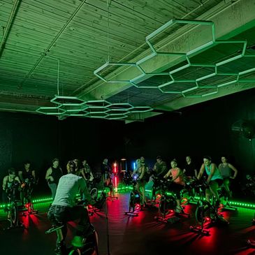 Indoor cycling class with neon green and red lights in a dark room.