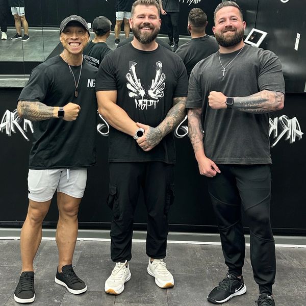 Three men posing together in a gym, smiling and showing off their muscular arms.