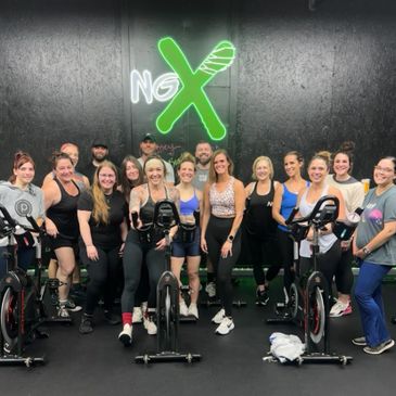 Group of people posing in a gym with exercise bikes and a neon sign.