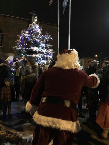 Santa Claus greeting people near a decorated Christmas tree at night.