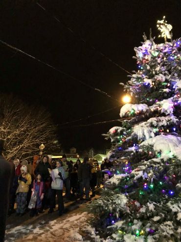 Snow-covered Christmas tree with colorful lights and a crowd gathered at night.