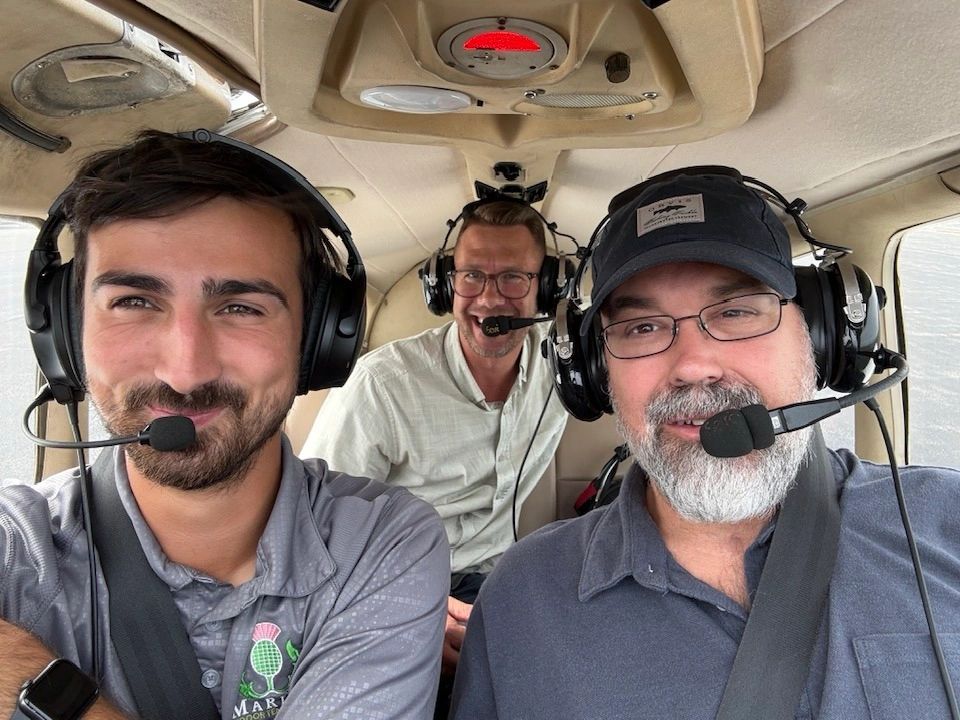 Three men with headsets inside a small aircraft, smiling at the camera.