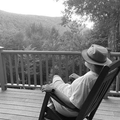 Person relaxing on a porch with a hat, overlooking a forested mountain landscape.