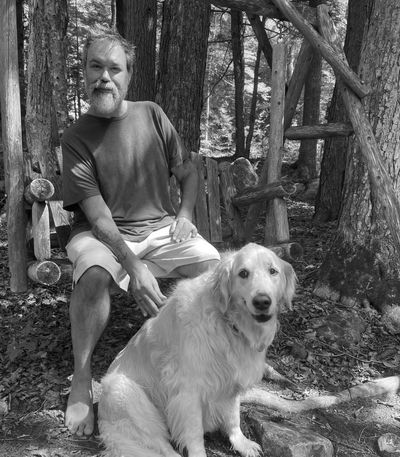 A man in a maroon shirt sitting barefoot on a rustic wooden bench with a golden retriever in a forest.
