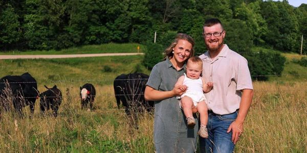 Happy family with baby standing in a grassy field with cows in the background.