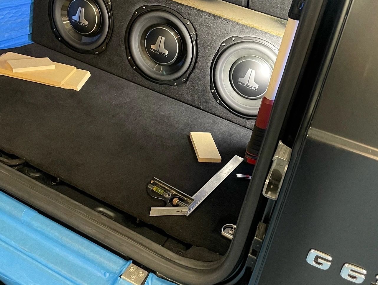 Three JL Audio subwoofers installed in a car trunk with measuring tools.