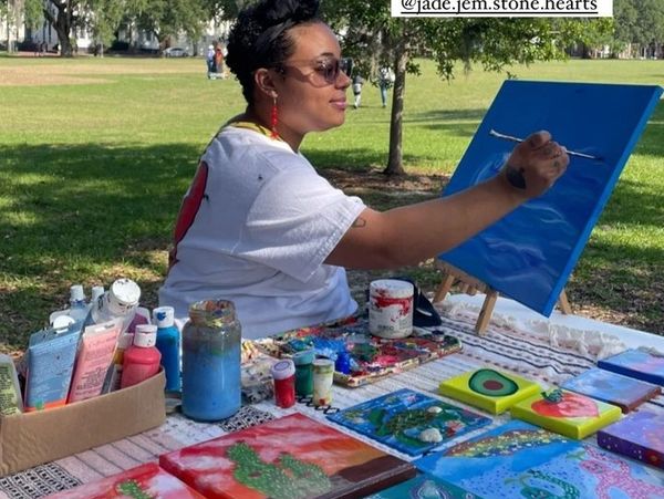 Artist painting a blue canvas outdoors with colorful artworks displayed.