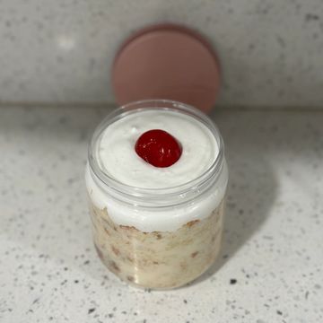 A jar of creamy dessert topped with a single red cherry on a speckled countertop.