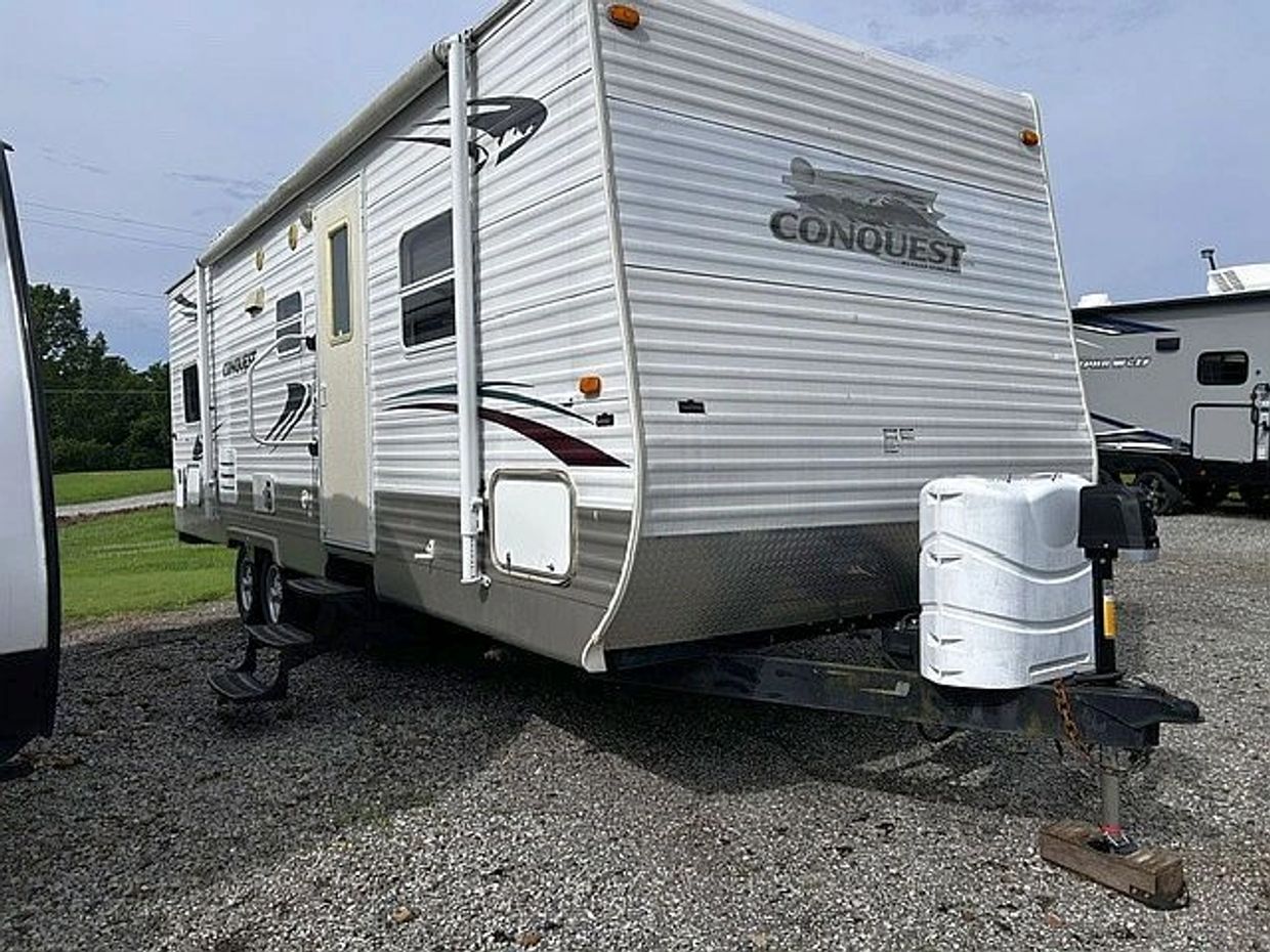 A white Conquest travel trailer parked on gravel with stairs extended.