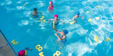 Children learning to swim in a pool with floating number markers.