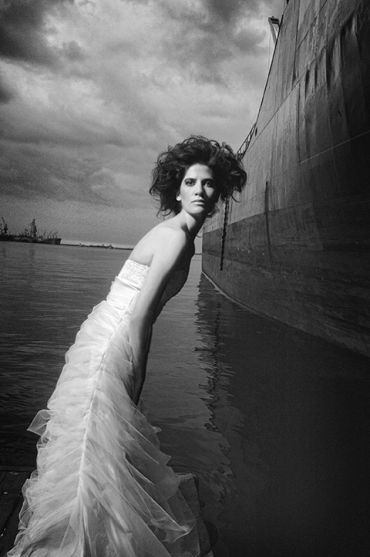 Woman in a flowing white dress near a large ship at a harbor.
